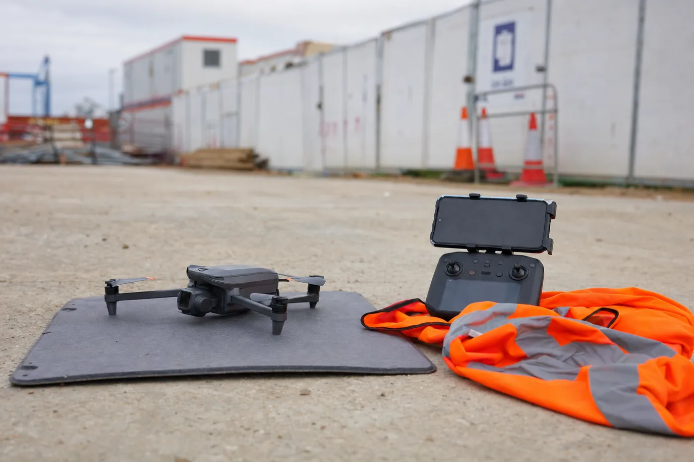 Folding camera drone on a launch pad beside a controller and hi-vis vest at the edge of a construction site