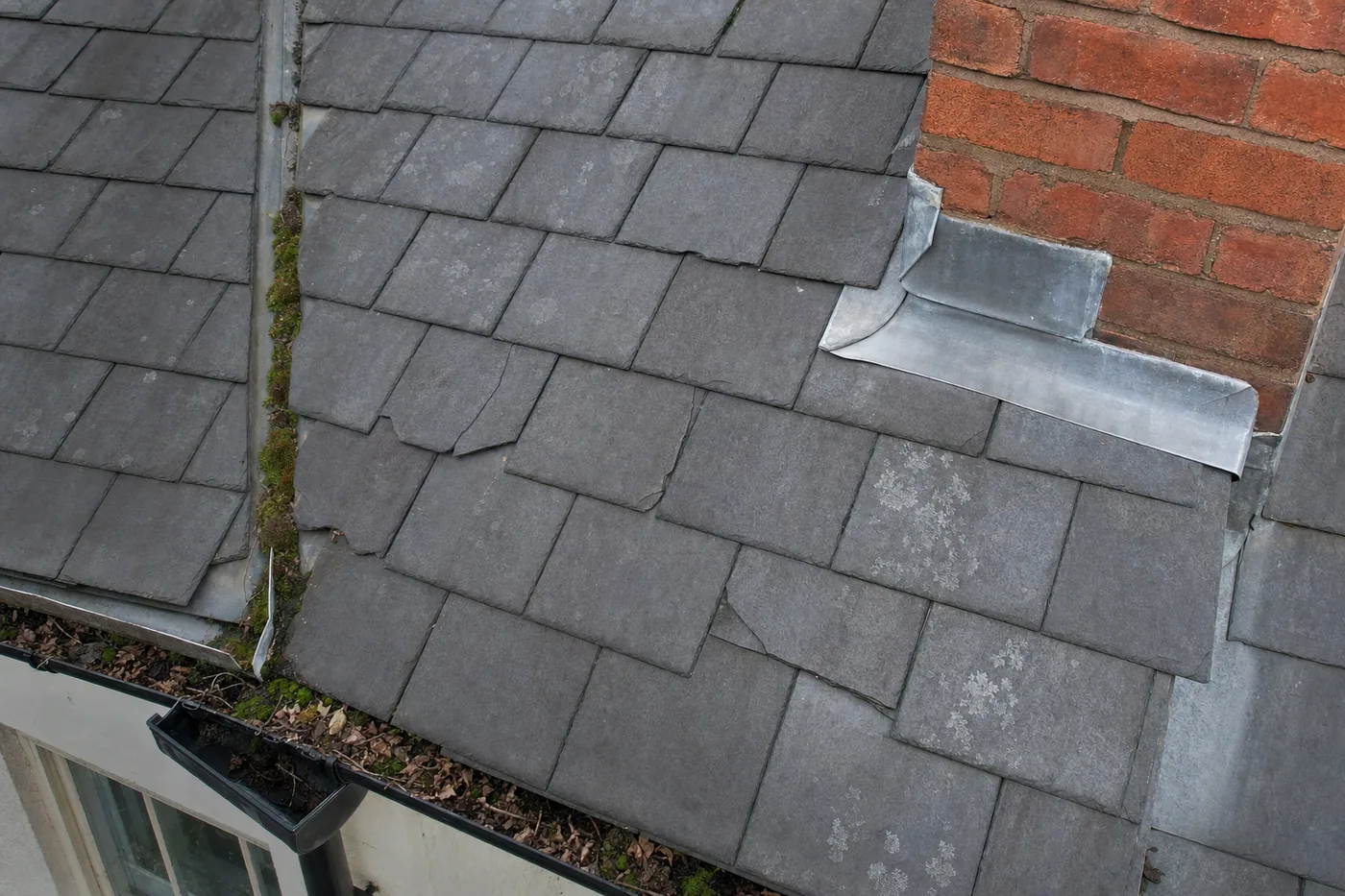 Close aerial view of a slate roof showing slipped tiles, moss in the valley and lead flashing at the chimney